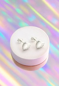 Hjärtformade silverörhängen med fem små klara accenter, vilande på en ljusrosa utställningsplatta mot en holografisk bakgrund.