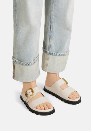 Sandalias de color beige claro con una correa ancha y una hebilla dorada, combinadas con pantalones de mezclilla azul claro, arremangados y con dobladillos deshilachados.