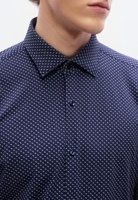 Μπλε πουκάμισο με κουμπιά και γεωμετρικό λευκό μοτίβο, με γιακά τύπου spread, κοντά μανίκια και κούμπωμα μπροστά με κουμπιά.