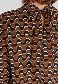 Mönstrad blus med en vågig design i olivgrön, svart, orange och krämfärgad. Bakdelen har en rosettknäppning vid halsringningen.