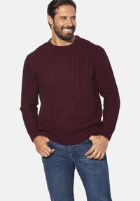 Bordeauxroter Strickpullover mit strukturiertem Zopfmuster, gerippten Bündchen und Saum. Kombiniert mit blauer Jeans.