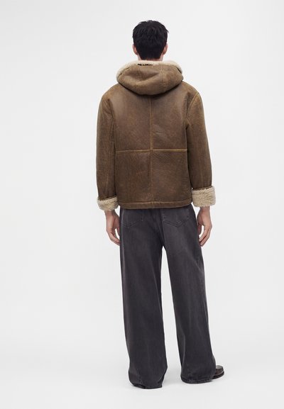 Man seen from behind wearing a brown shearling jacket with hood and cuffs, paired with loose dark gray pants and dark shoes.