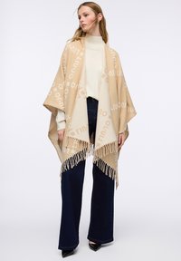 Poncho beige intrecciato con grande motivo logo, rifinito con frange. Indossato sopra un maglione chiaro e jeans svasati scuri.