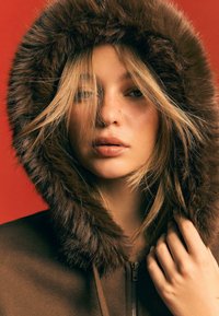 Bruine hoodie met bontvoering en een volledige capuchon, met een rits en textuurstof. Het model heeft een natuurlijke make-up en een rommelige haarstijl voor een neutrale achtergrond.