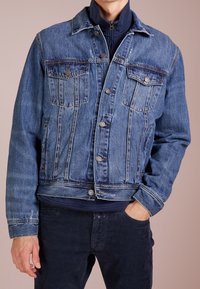 Muž v modré džínové bundě přes námořnický zipový svetr, s jednou rukou v kapse, stojí na pozadí v jednobarevné béžové barvě.