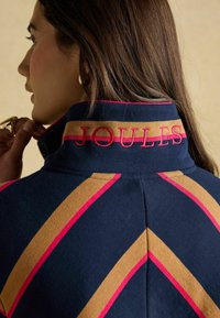 Jachetă marină cu dungi diagonale în aurie și roz, având un guler înalt și cu broderie "JOULES" pe spate. Textură moale.