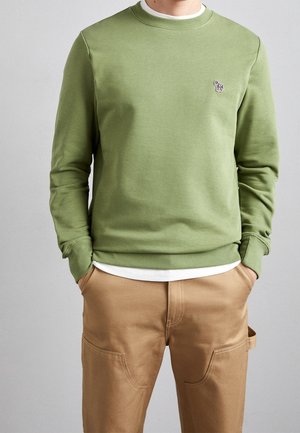 Mand iført olivengrøn sweatshirt med lille zebra-logo på brystet og beige bukser med hænderne i lommerne mod grå baggrund.