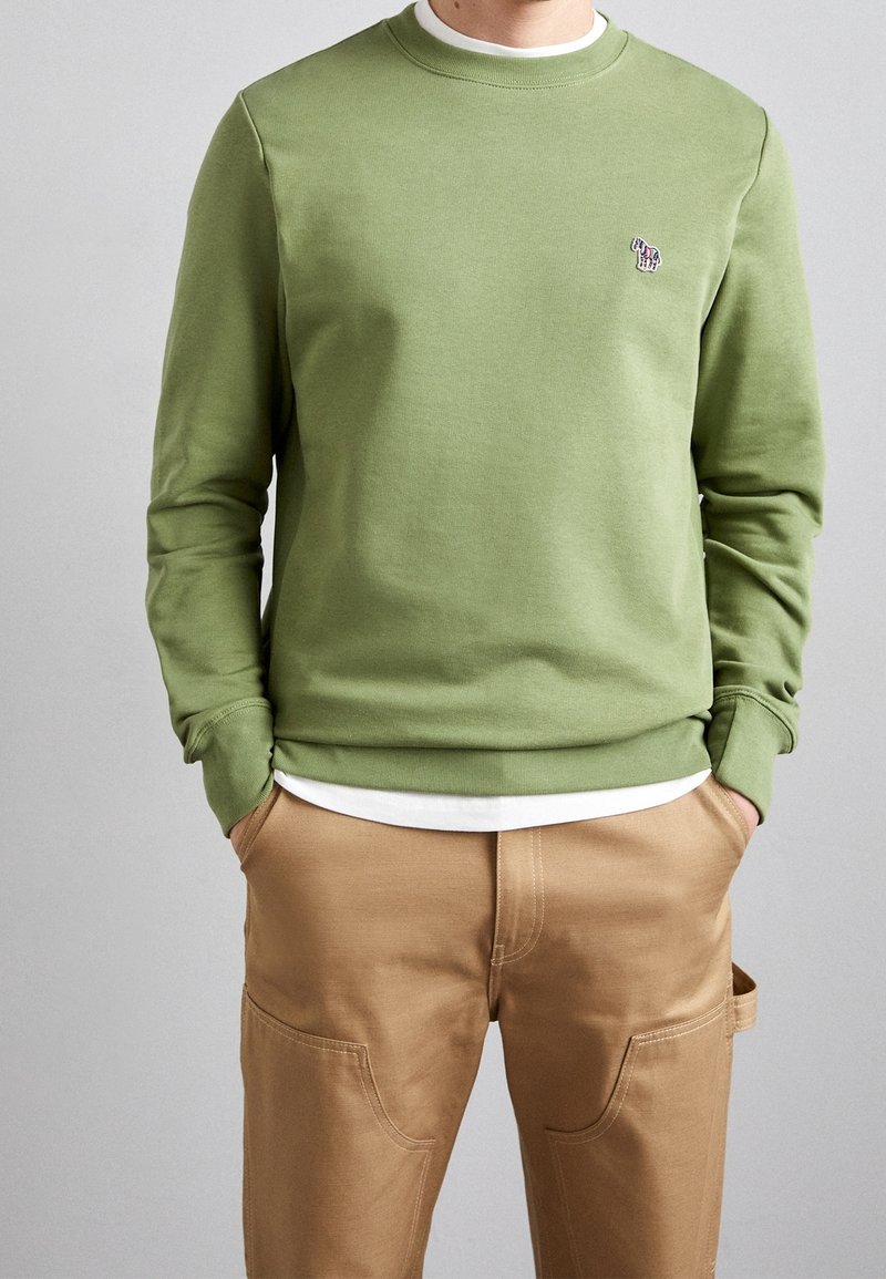 Homme portant un sweat-shirt vert olive avec un petit logo zèbre sur la poitrine et un pantalon beige, les mains dans les poches, sur fond gris.