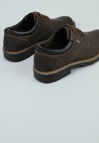 Zapatos de cuero marrón con cordones, suelas negras robustas y costuras visibles, colocados sobre una superficie gris claro.