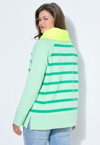 Mujer con cabello largo y castaño que lleva una sudadera con capucha verde menta con rayas verde oscuro y una capucha de color amarillo neón, vuelta para mostrar la parte trasera.