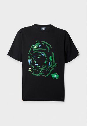 Homme portant un t-shirt noir à motif, un jean large gris clair et des chaussures noires et blanches, debout les mains dans les poches.