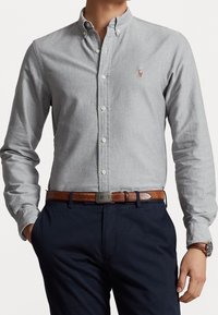 Uomo che indossa una camicia grigia chiaro con bottoni infilata in un pantalone blu navy, con cintura in pelle marrone e orologio da polso, mani rilassate lungo i fianchi.