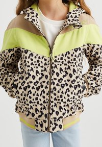 Bambino con giacca a stampa leopardata, con blocchi di colore beige, verde e marrone, zip frontale e mani nelle tasche.