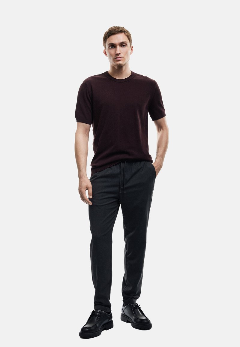 Donker kastanjebruin trui met korte mouwen, gecombineerd met grijze broek met trekkoord en zwarte leren schoenen. Effen kleuren, eenvoudig ontwerp, lichtgewicht stof.