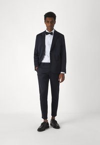 Tumed ülikond slim fit'iga, millel on ühe nupuga bleiser ja kitsad püksid. Kantakse koos valge särgi ja mustade kleitkingadega.