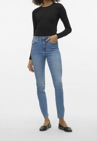 Frau in hochgeschnittenen hellblauen Skinny-Jeans, schwarzem Langarmoberteil und schwarzen Ballerinas, die mit einer Hand in der Tasche steht.