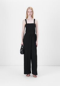 Mono negro con top fruncido, tirantes finos, silueta de pierna ancha, tela texturizada, combinado con un pequeño bolso negro y sandalias planas.
