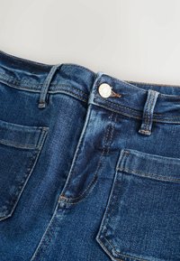 Tmavě modré džíny s vysokým pasem, předním zapínáním na knoflík a dvěma zadními kapsami. Vyznačují se kontrastním prošitím a hladkou texturou.