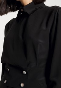 Černá blůza s strukturovaným límcem, saténovou texturou a knoflíky ve tvaru perel. Obsahuje precizní detaily a jemně vyšívané logo.