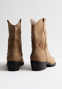 Botas de tobillo de ante marrón con un bajo tacón negro y cortes curvados en el tobillo, que muestran una textura suave y un diseño sencillo.