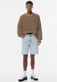 Braunes, strukturiertes Langarmhemd, hellblaue Jeansshorts, schwarze Slipper mit weißen Akzenten, weiße Socken, geschichtete Kettenhalsschmuck.