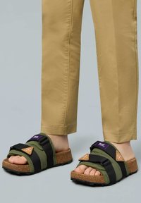 Green and black sandals with cork footbed, adjustable straps, and tan accents. Textured sole provides grip, featuring purple label on strap.