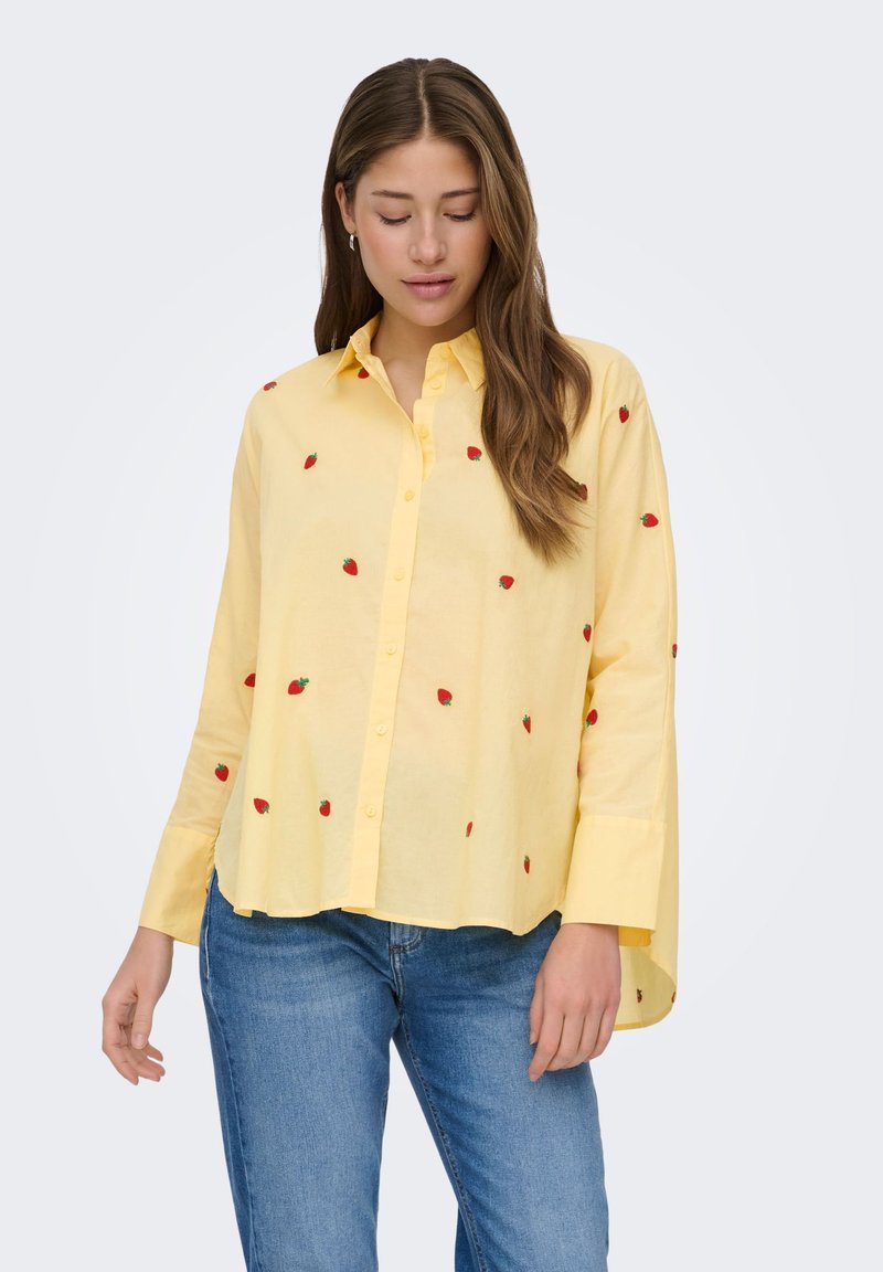 Femme portant une chemise jaune ample à boutons avec de petits motifs de fraises rouges, associée à un jean bleu, debout devant un fond uni.