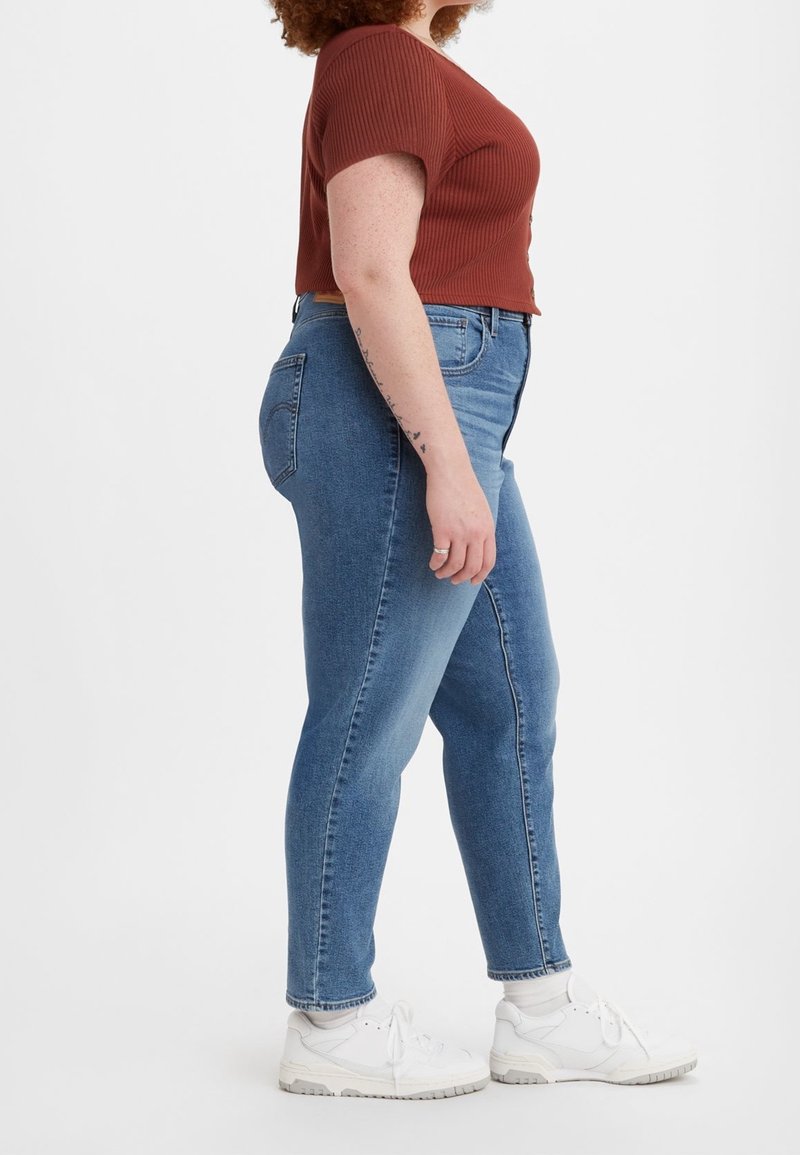 Personne aux cheveux bouclés portant un haut à manches courtes couleur rouille, un jean bleu taille haute, des chaussettes blanches et des baskets blanches, debout de profil.