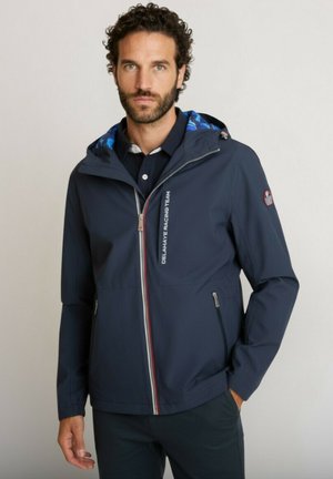 Homme portant une veste zippée à capuche bleu marine avec le texte « Delahaye Racing Team » et deux poches zippées, debout devant un fond uni.