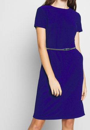 Kobieta ubrana w królewsko niebieską sukienkę o długości do kolan i krótkim rękawie, z cienkim srebrnym paskiem, stojąca z jedną ręką w kieszeni.