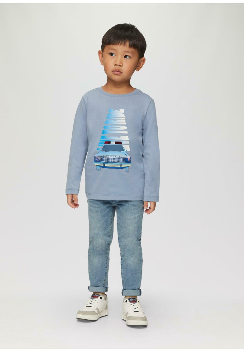 Langärmliger blauer Pullover mit einem Polizeiauto-Design, kombiniert mit hellblauen Jeans und weißen Turnschuhen mit schwarzen Akzenten.