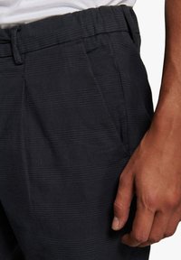Pantalones a cuadros gris oscuro con una tela texturizada, con un frente plano, pliegues y una cintura elástica. Una mano descansa en el bolsillo lateral.