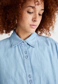 Lyseblå denimskjorte med klassisk krage, knappeåpning og et lett teksturert stoff. Har synlige sting og runde knapper.
