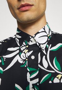 Chemise noire présentant un motif floral avec des fleurs blanches et des feuilles vertes. Design à col avec des boutons blancs sur le devant.