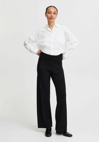 Witte blouse met lange mouwen, gecombineerd met zwarte wijde broek en zwarte schoenen. Eenvoudig, op maat gemaakt ontwerp met een relaxte pasvorm.