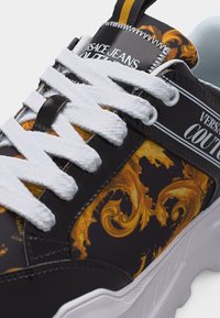 Μαύρο και χρυσό αθλητικό παπούτσι Versace Jeans Couture με λευκά κορδόνια και περίτεχνο μοτίβο μπαρόκ στο πλάι και στη γλώσσα.