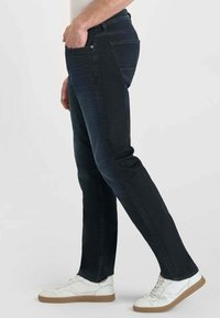 Jean slim fit bleu foncé en tissu denim avec une texture lisse. Doté de cinq poches et d'une taille classique. Porté avec des baskets blanches.