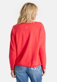Frau mit blonden Haaren, die einen roten Sweatshirt mit dem Text "enjoy the now" auf dem Rücken und eine blaue Jeans mit einem weißen Design auf der Tasche trägt.