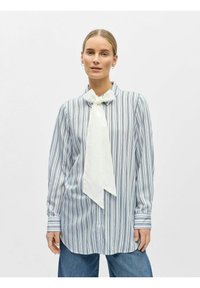 Femme portant un chemisier à manches longues bleu et blanc à rayures avec un grand noeud blanc, associé à un jean bleu, debout devant un fond uni.