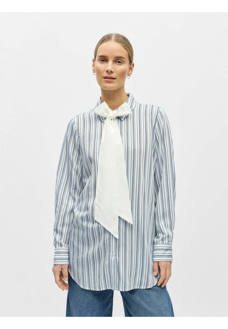 Femme portant un chemisier à manches longues bleu et blanc à rayures avec un grand noeud blanc, associé à un jean bleu, debout devant un fond uni.