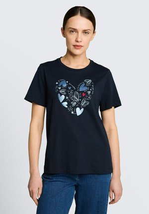 Kobieta z ciemnymi, zaczesanymi do tyłu włosami nosi granatowy t-shirt z różnymi niebieskimi i czerwonymi motywami serc tworzącymi większy kształt serca, w połączeniu z niebieskimi dżinsami.
