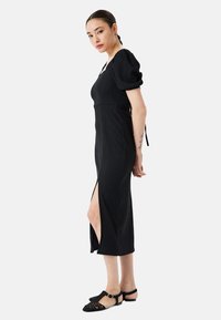 Vestido negro de longitud a la rodilla con mangas abullonadas, tejido texturizado y aberturas laterales. Se lleva con zapatos planos negros de diseño trenzado y correa en el tobillo.