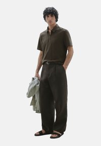 Polo shirt corto marrone scuro con colletto a zip, abbinato a pantaloni scuri larghi. Il modello tiene in mano una giacca grigia chiara e indossa sandali neri.