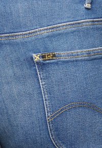 Los jeans de denim azul presentan un bolsillo trasero con costuras doradas y una pequeña etiqueta negra con la marca "Lee". Tejido texturizado con un ligero desgaste.