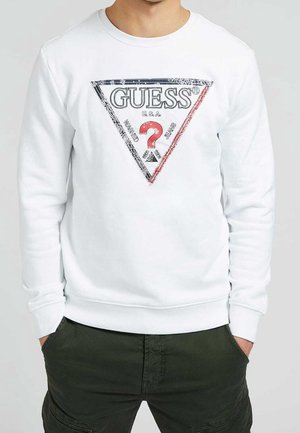 Camisa blanca de manga larga con un gran logo triangular de GUESS que incluye un signo de interrogación rojo y texto en negro, rojo y blanco, combinada con pantalones verde oscuro.