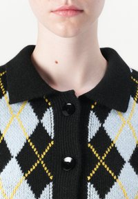 Pletený cardigan v černé, světle modré a žluté argyle vzoru. Disponuje černým límcem a dvěma velkými černými knoflíky vpředu.