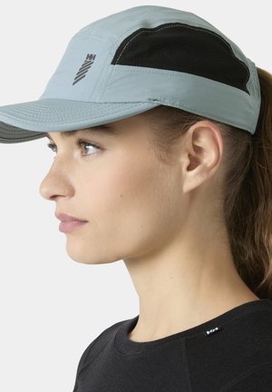 Mujer con gorra deportiva azul clara y negra y camiseta negra de cuello redondo, mirando hacia la izquierda sobre un fondo liso.