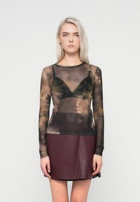 Μακρυμάνικο τοπ από διχτυωτό ύφασμα με καφέ και μαύρο tie-dye σχέδιο, φορτωμένο πάνω από ένα μαύρο bralette, συνδυασμένο με μια μπορντό δερμάτινη μίνι φούστα.