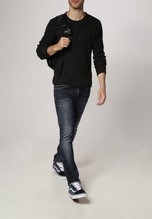 Uomo che cammina indossando un maglione nero, jeans scuri, sneakers blu navy e portando uno zaino nero su una spalla contro uno sfondo semplice.