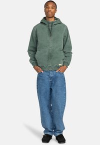 Sudadera verde de algodón con cremallera en el frente, capucha con cordón y bolsillos laterales, combinada con jeans de mezclilla azul holgados y zapatos negros.